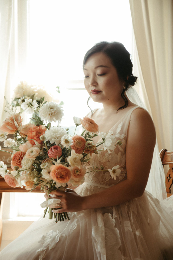 A bride looking at her bouquet of white and peach colored flowers.