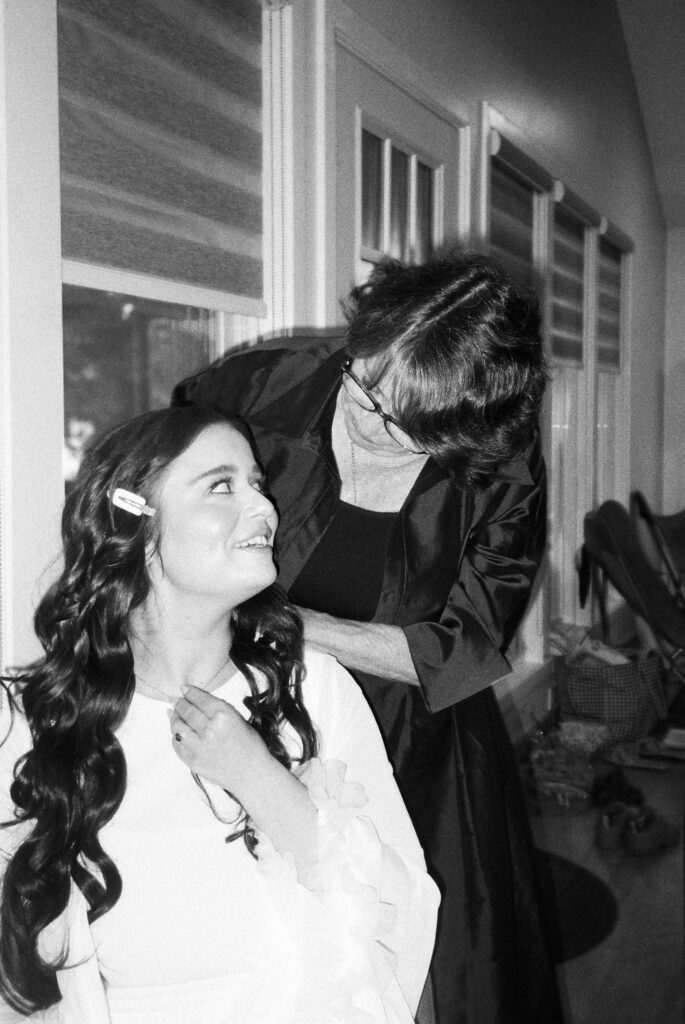 Candid moment of the bride's future mother in law putting a necklace on her, shot on black and white film.