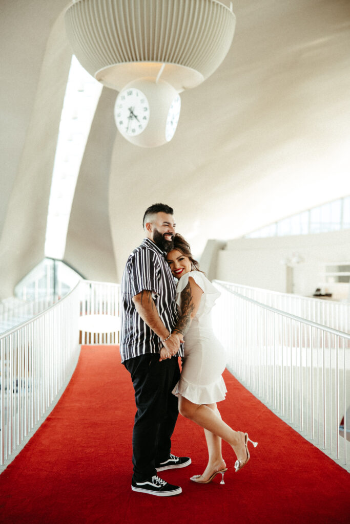 Engaged couple posing in the TWA Hotel’s bold red lounge, surrounded by sleek architecture.