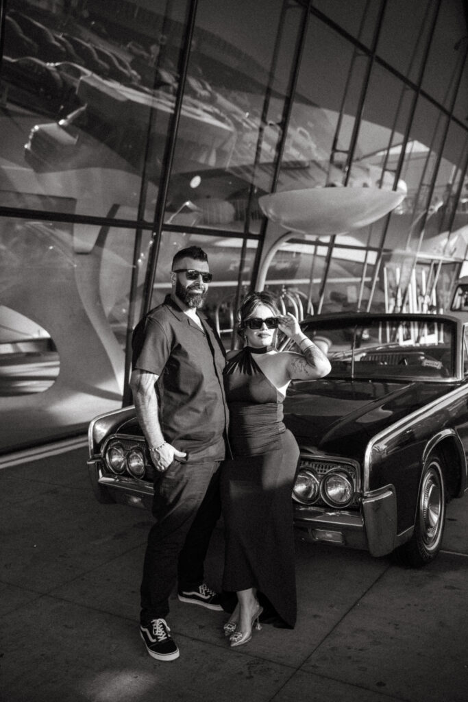 Engaged couple posing next to a vintage car parked in front of the TWA Hotel.