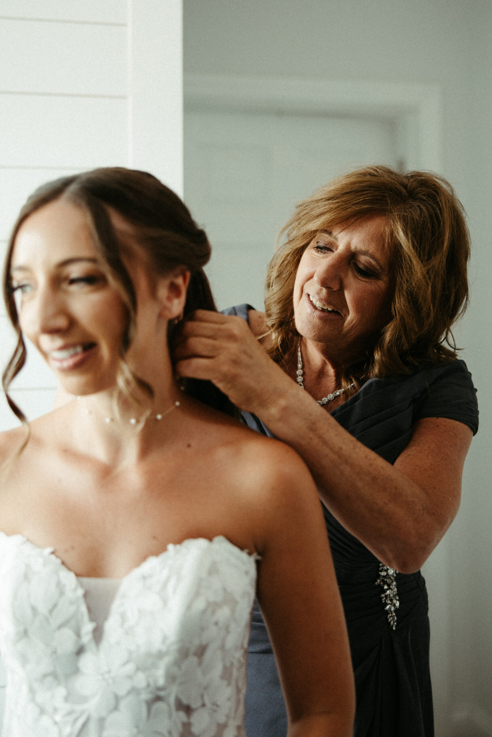 A mother smiles with joy as she fastens her daugher's, the bride, necklace on her wedding day.