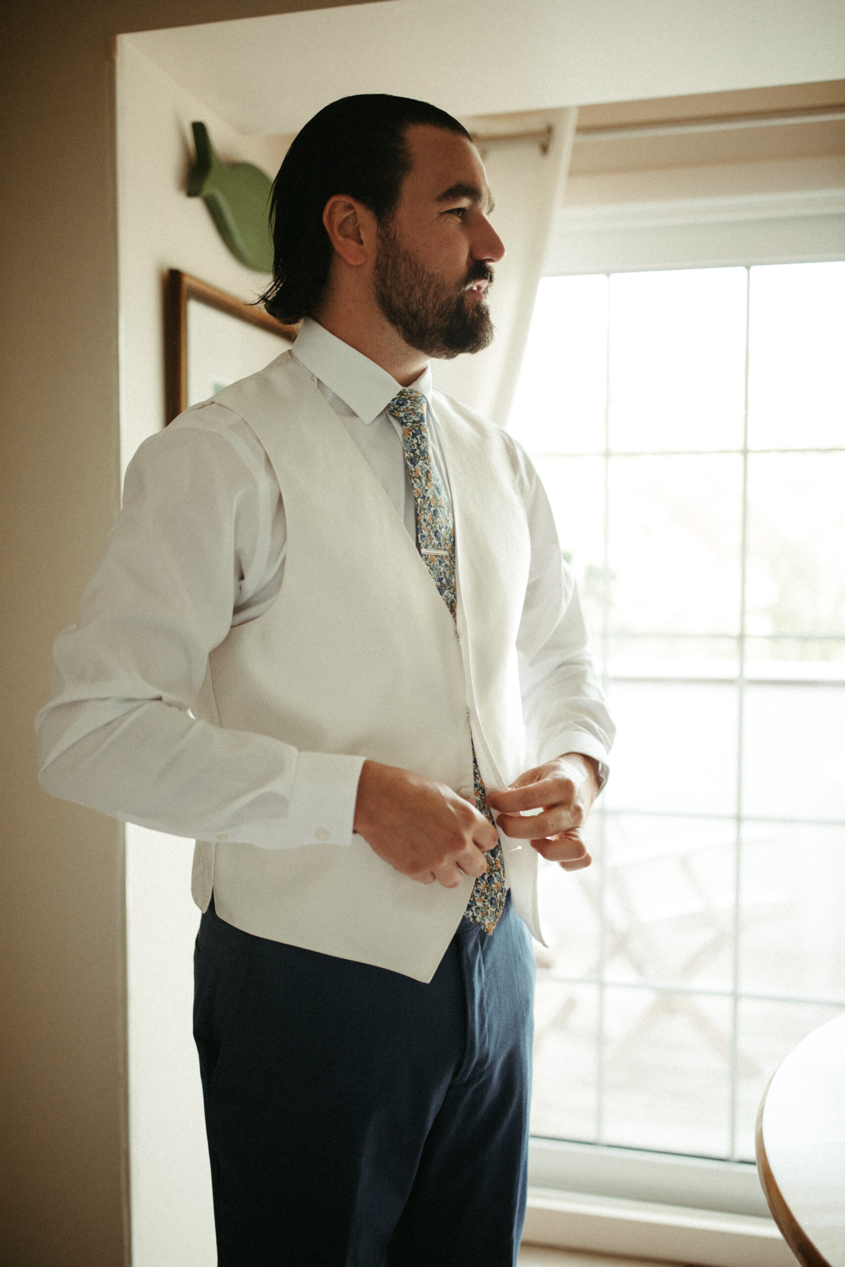 A groom in a tailored suit buttons his vest in front of a window.