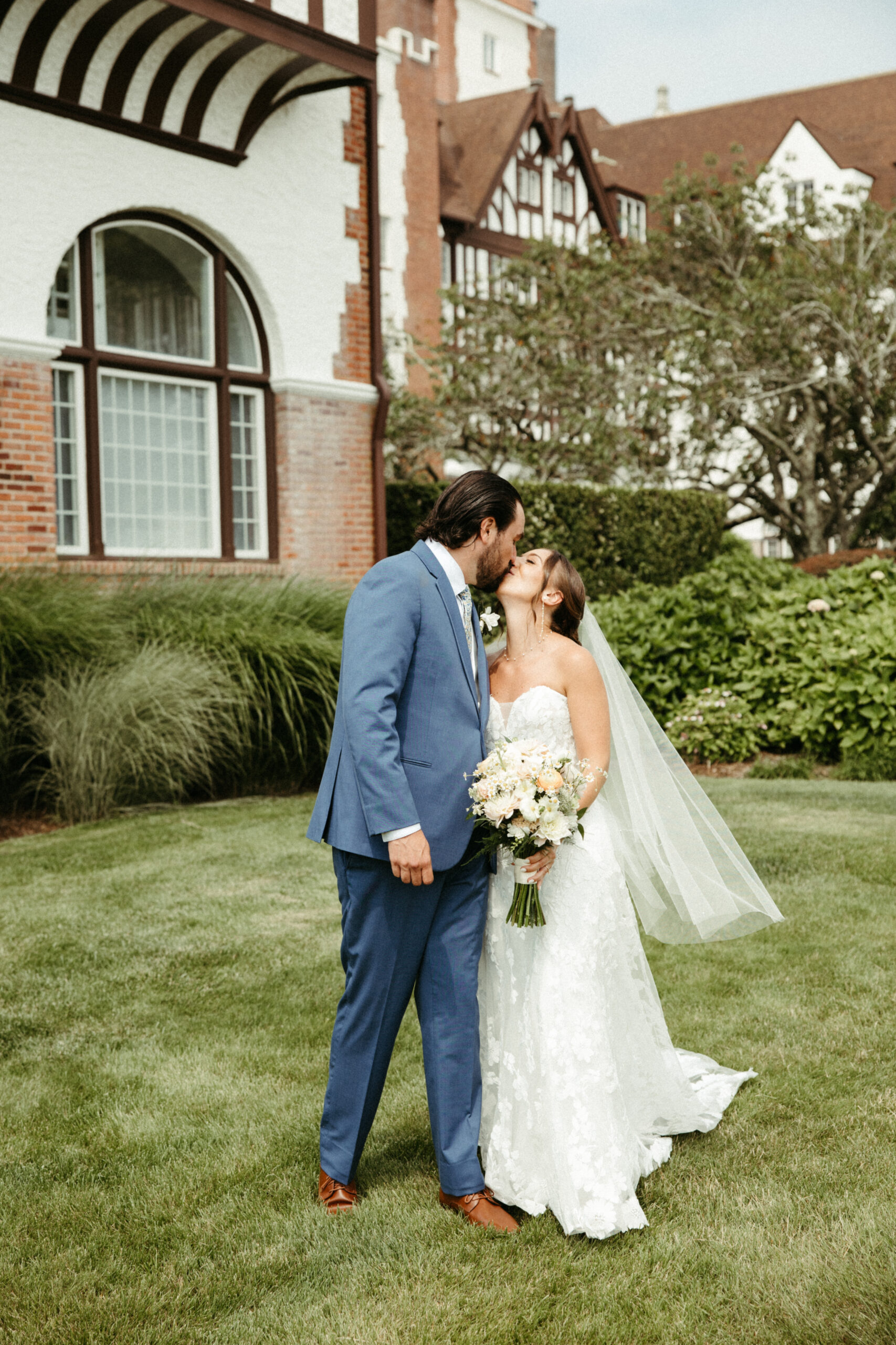 The couple stands hand-in-hand and kiss in front of the grand Montauk Manor, the historic architecture framing their love.