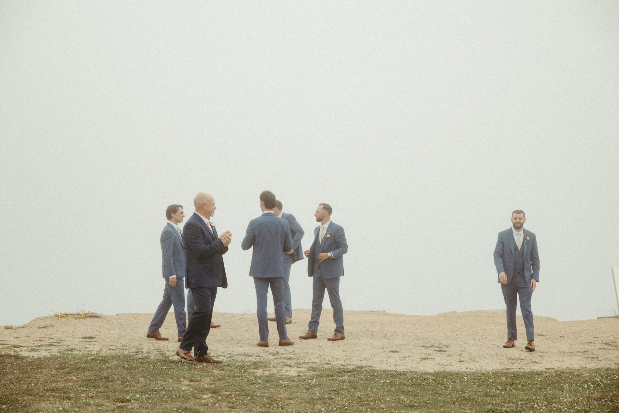 Guests dressed in elegant attire stand near the cliff’s edge, gazing at the misty ocean beyond.