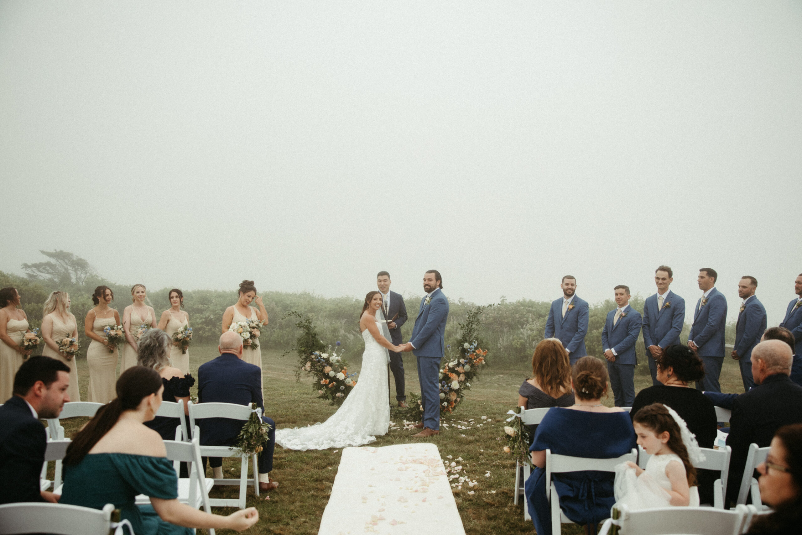  Rows of wooden chairs face the cliff’s edge, where the couple stands beneath a floral-adorned arch with the lighthouse faintly visible.