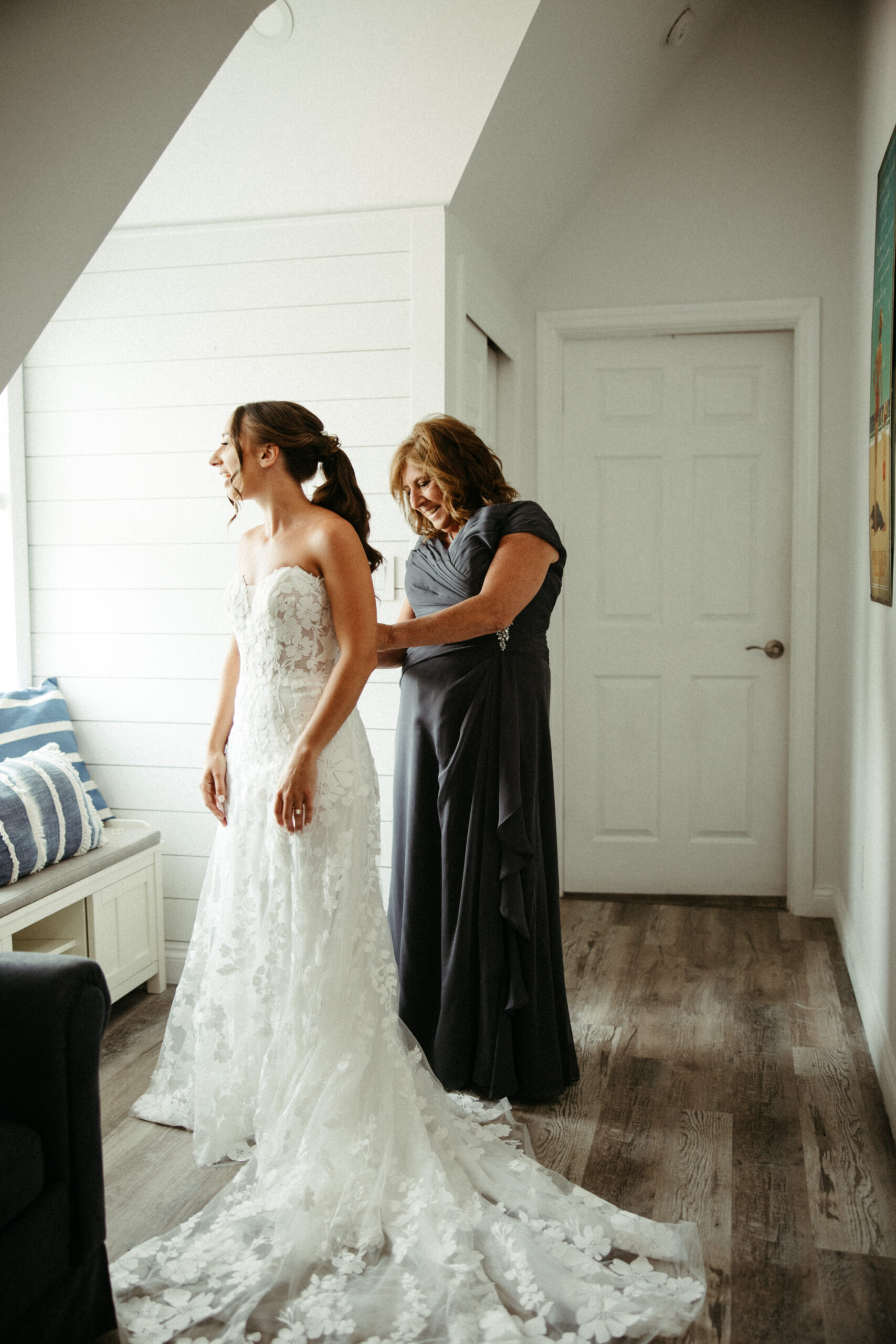 A mother buttons her daughters wedding dress bathed in the morning light streaming through a window.