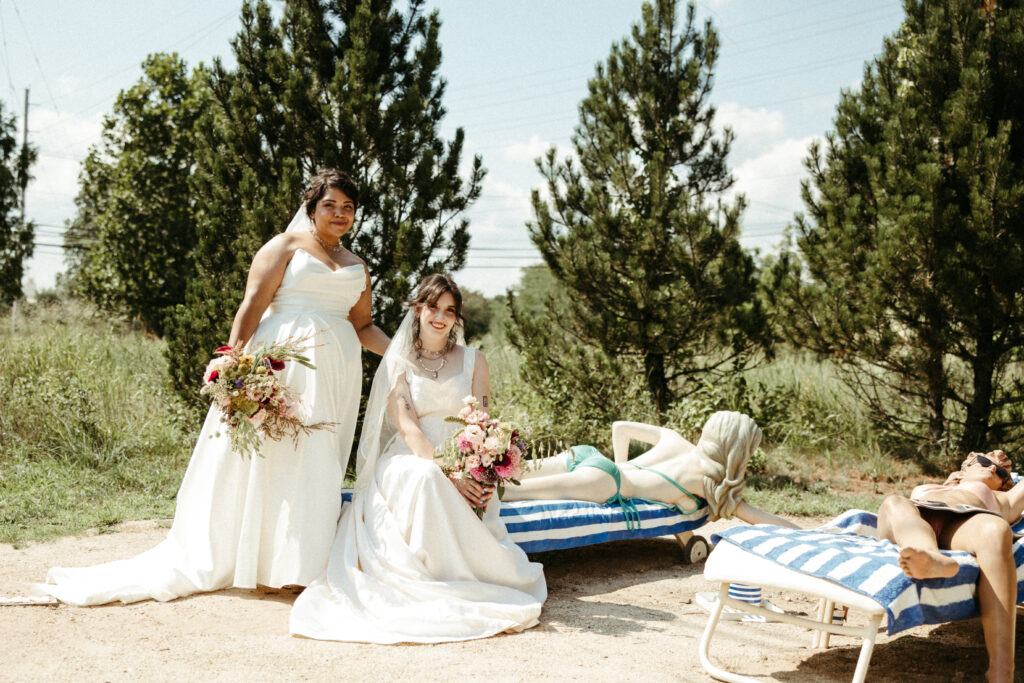 A lesbian couple on their wedding day pose with an art installation of the two women sunbathing at the Grounds For Sculpture.