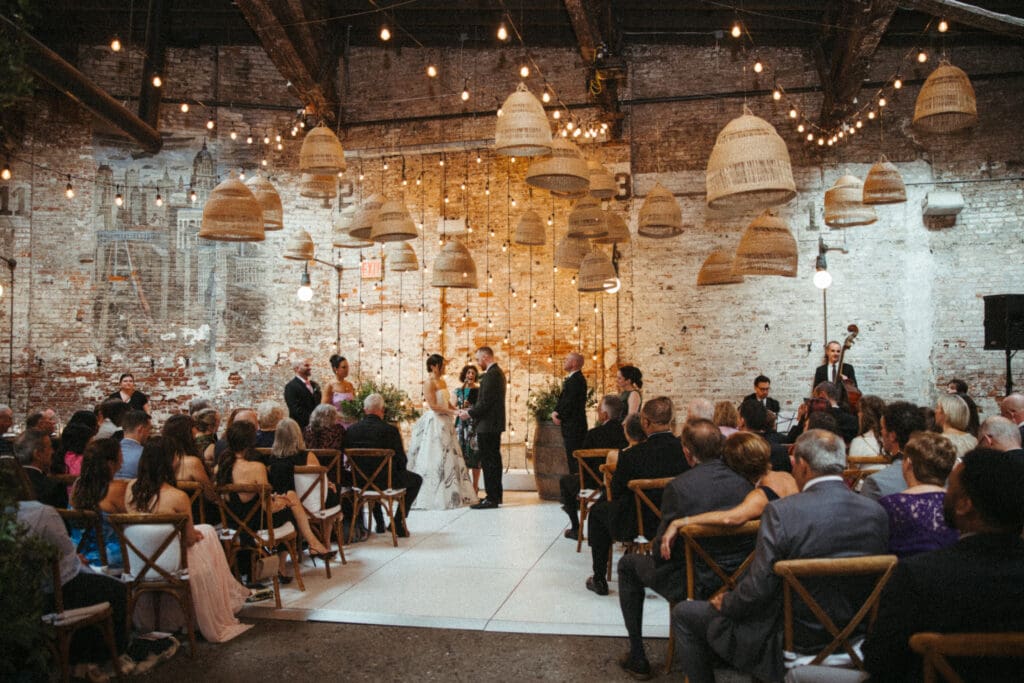Wide view of Adrianna and Jack’s Houston Hall wedding ceremony with hanging bistro lights, exposed brick, and guests seated on both sides
