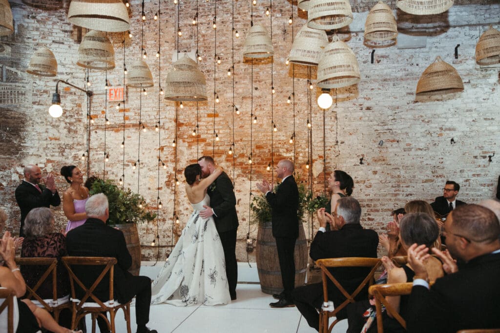 Adrianna and Jack sharing their first kiss during their Houston Hall wedding ceremony under glowing string lights