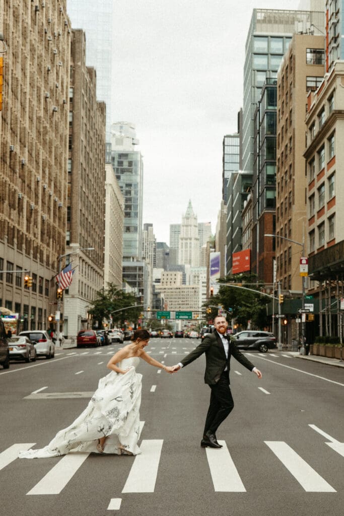Bride and groom holding hands while crossing Houston Street in New York City with the skyline behind them