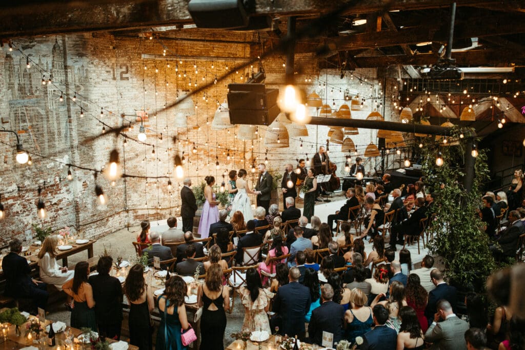 Overhead view of Houston Hall wedding ceremony with Adrianna and Jack exchanging vows beneath glowing string lights and woven pendants