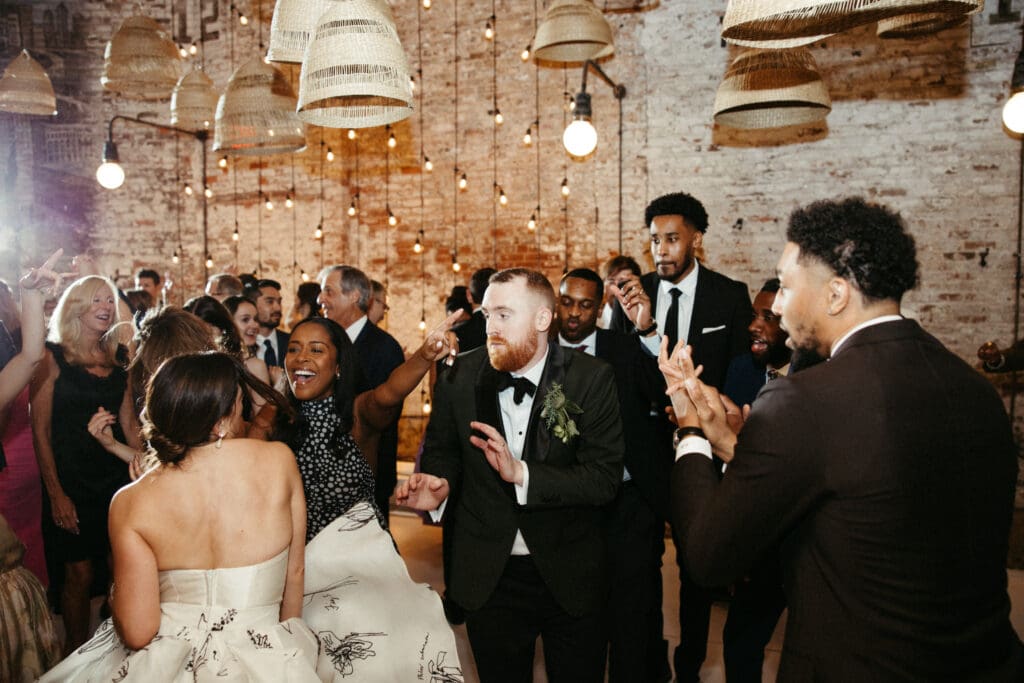 Bride and groom laughing together on the dance floor at their Houston Hall wedding as guests dance under string lights in the historic NYC venue