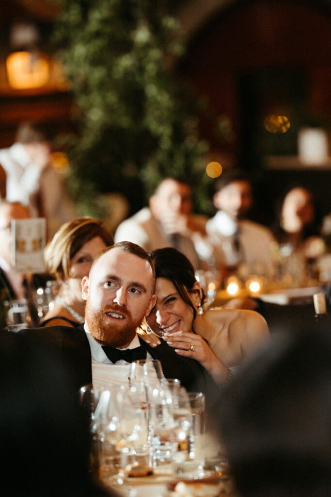 Bride rests her head on her groom’s shoulder during heartfelt wedding speeches inside Houston Hall, surrounded by candlelight and guests.