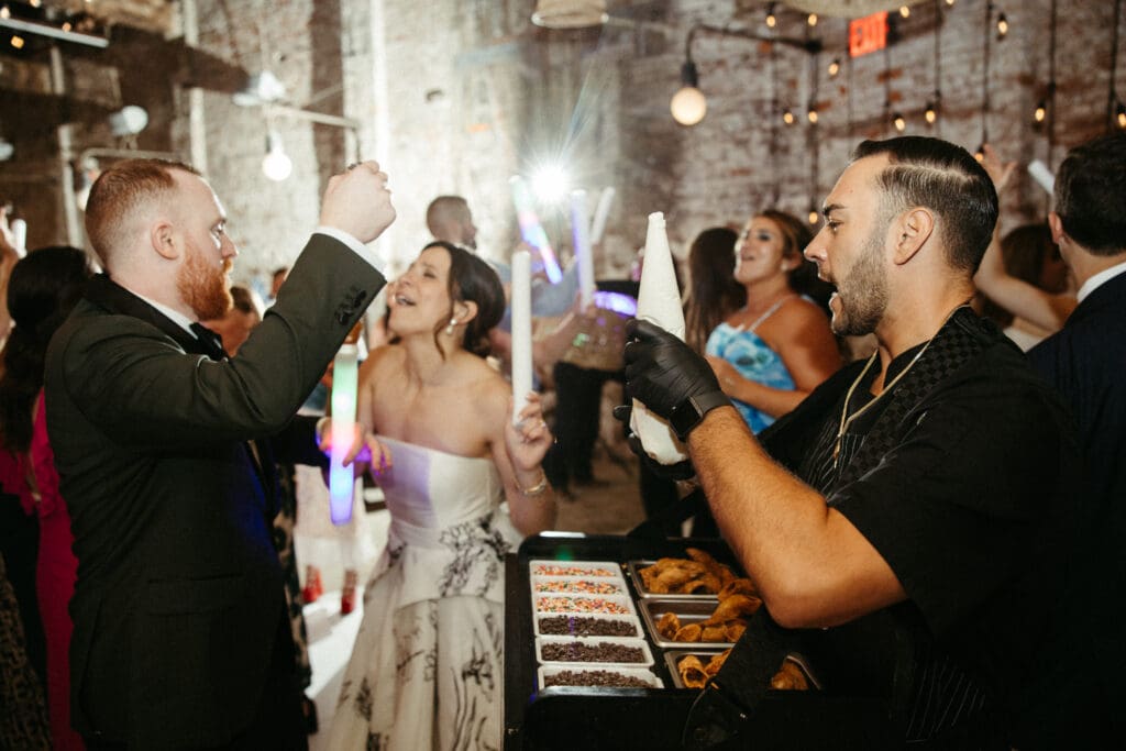 Bride and groom laugh as the Cannoli Guy serves fresh cannolis on the dance floor at their Houston Hall wedding in New York City.