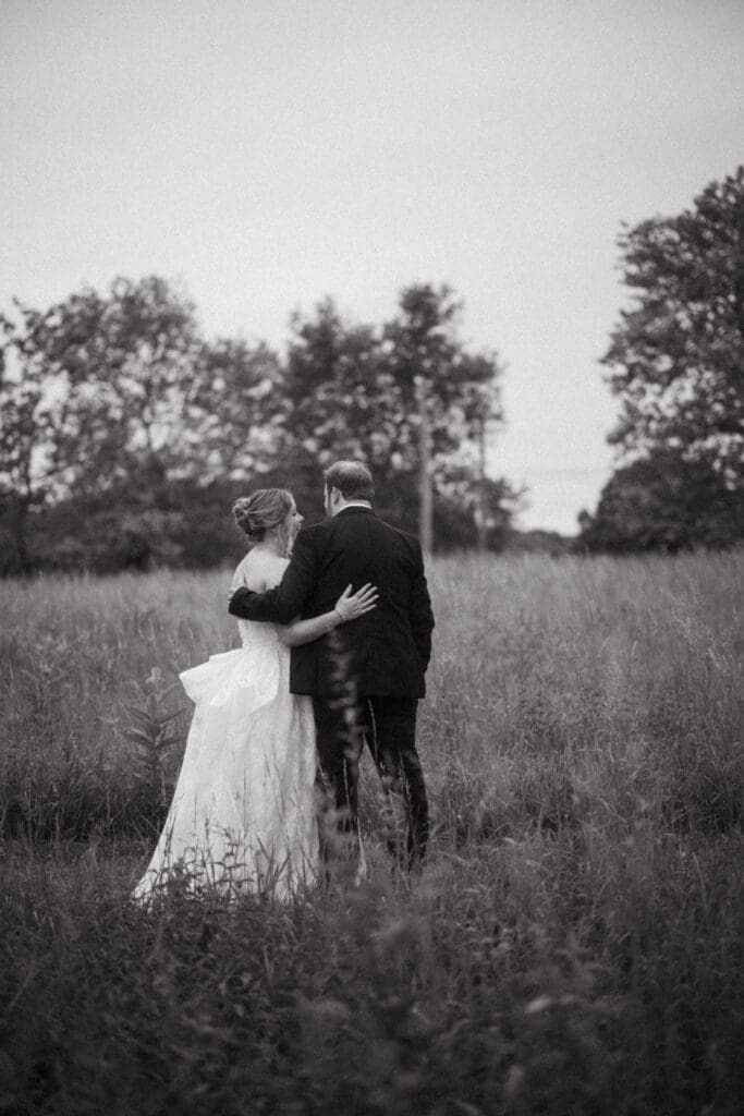 Bride and groom walking arm in arm through tall grass at Stone Acres Farm, captured in a romantic black and white wedding portrait.