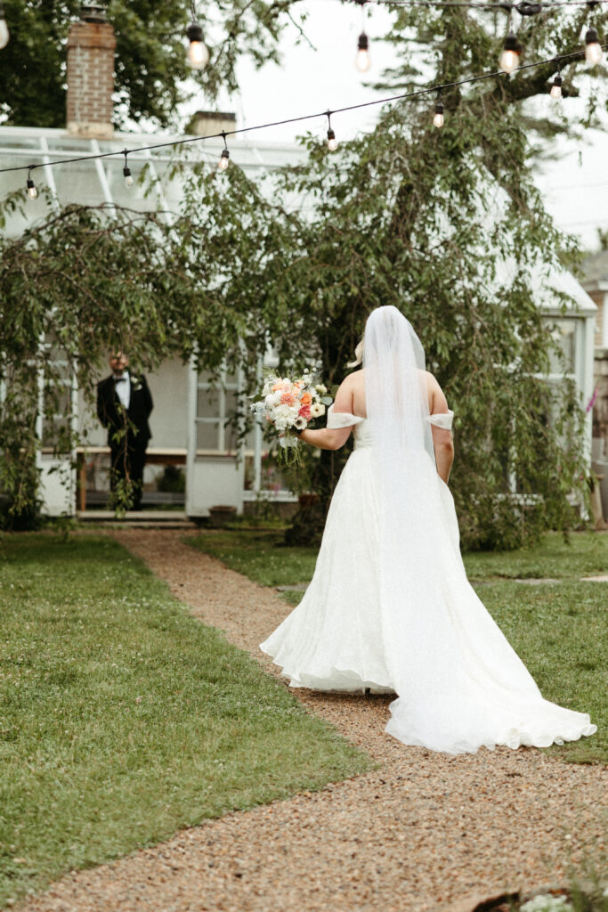 Bride walks down a garden path toward her groom for a private first look outside the greenhouse at Stone Acres Farm