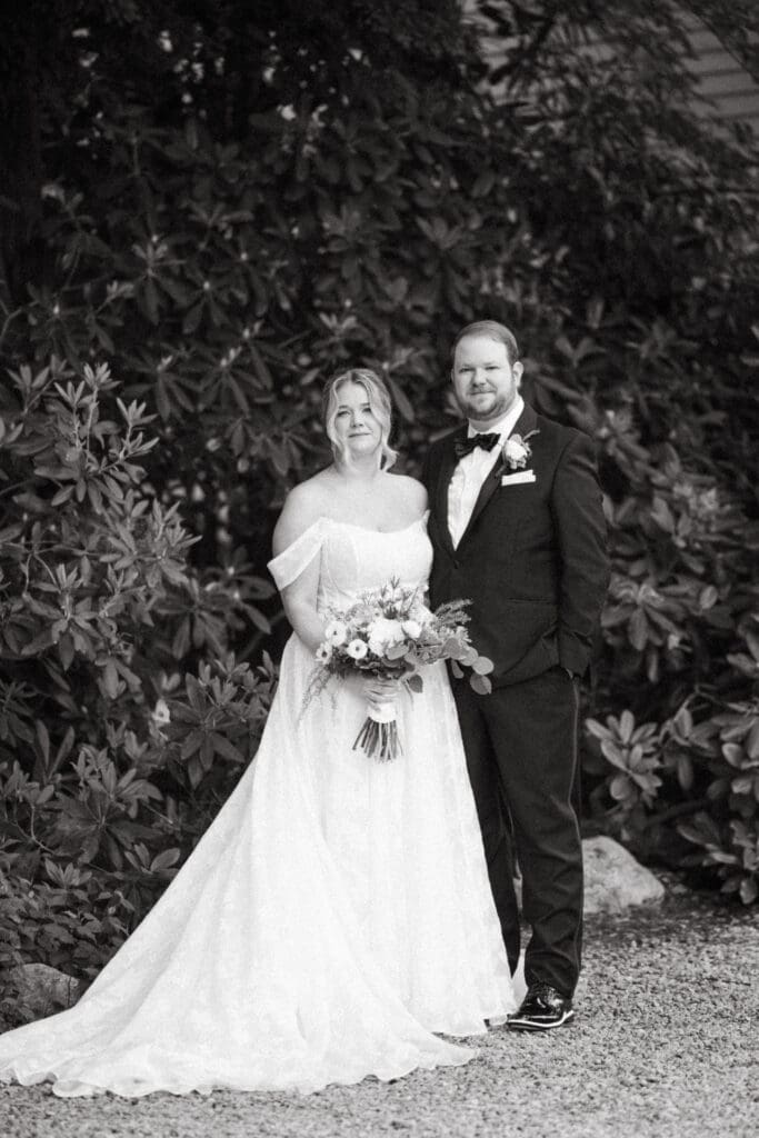 Bride and groom posing together with bouquet in front of lush garden foliage at Stone Acres Farm, photographed in elegant black and white.