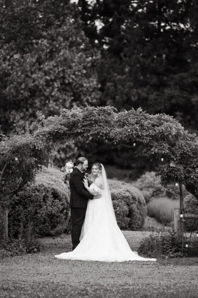 Bride and groom standing together beneath a garden arch at Stone Acres Farm, framed by greenery in a classic black and white wedding portrait.