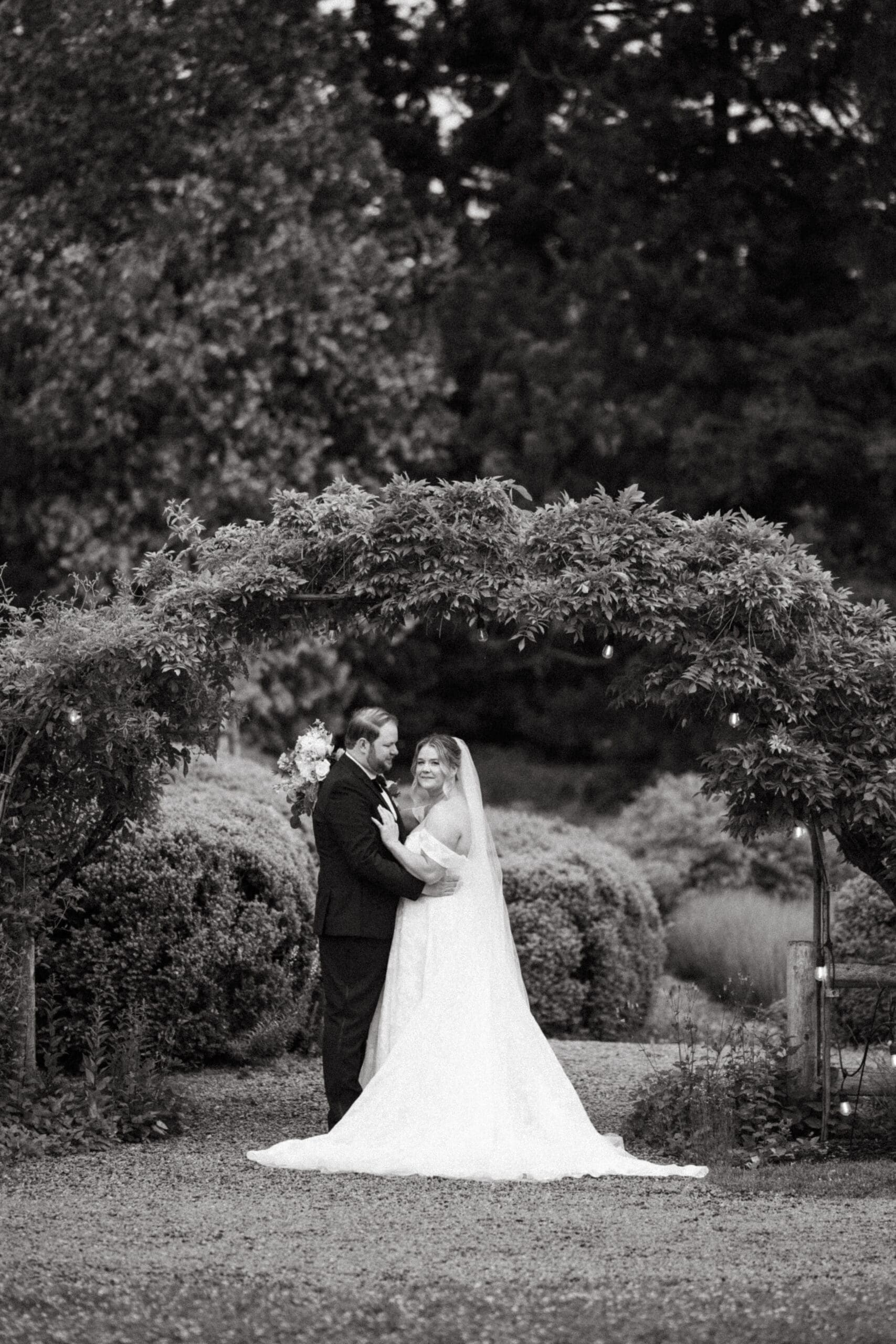 Bride and groom standing together beneath a garden arch at Stone Acres Farm, framed by greenery in a classic black and white wedding portrait.