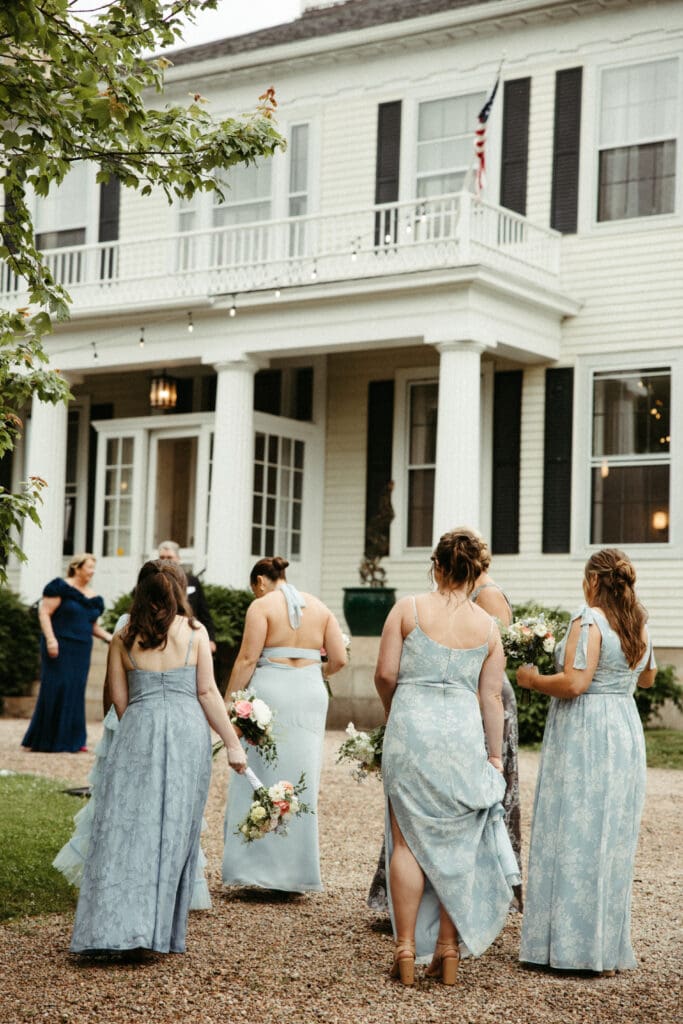 Bridesmaids in soft blue dresses walking toward the historic farmhouse at Stone Acres Farm in Stonington, Connecticut during a Stone Acres Farm wedding