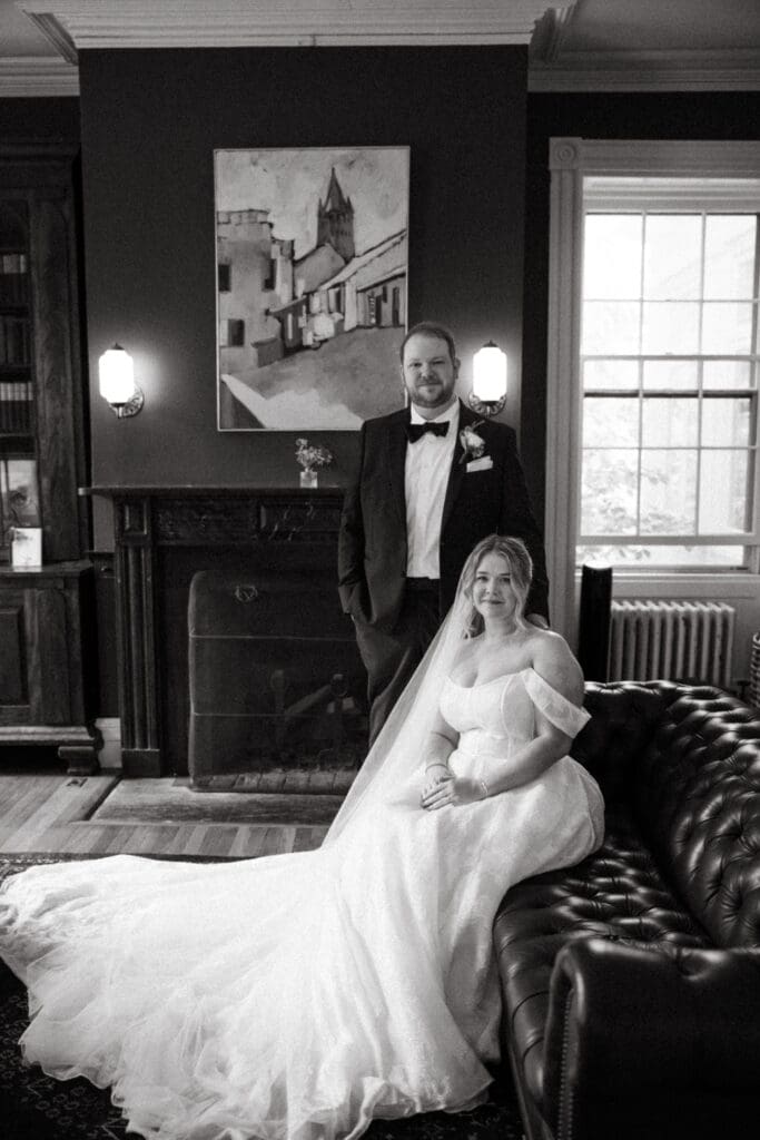 Bride seated on a leather sofa with groom standing behind her inside the historic Stone Acres Farm estate, photographed in timeless black and white.