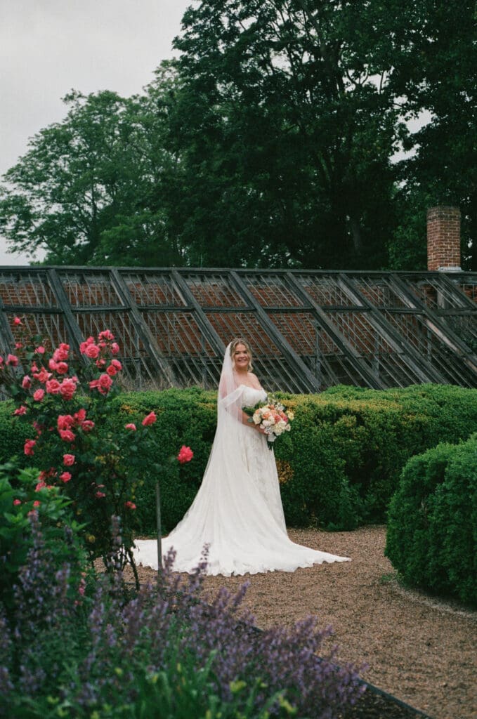 Bride standing in front of the historic Stone Acres Farm greenhouse surrounded by roses, captured on film in soft, overcast light.